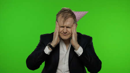 Portrait of drunk young clerk man with a hangover having headache. Emotions. Disheveled guy businessman in suit and festive cap looks at the camera. Chroma key. Office birthday party conceptの写真素材