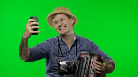 Portrait of man tourist with suitcase in hand is making selfies using mobile phone. Smartphone communication. Handsome man in blue shirt and hat with retro camera. Chroma key. Green screenの写真素材