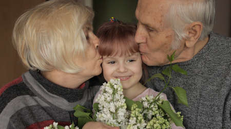 Happy senior grandfather and grandmother couple kissing on head their kid with flowers granddaughter at home. Senior lifestyle family concept. Family time relax with young child girl. Slow motionの写真素材