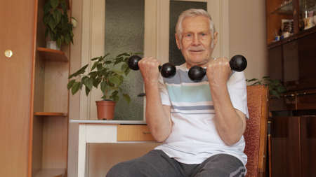 Senior elderly caucasian man in sportswear sitting on chair in living room, doing weight lifting dumbbell exercising at home. Active old retiree exercising in the morning. Leisure healthy lifestyleの写真素材