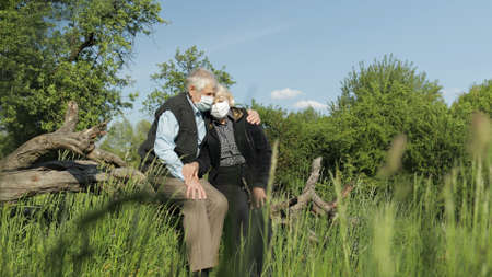 Senior old grandparents couple in medical masks sitting in park during COVID-19 coronavirus...