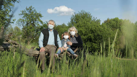 Grandparents with granddaughter together sitting in spring park in medical masks. Coronavirus covid-19 pandemia quarantine. Family time together. Grandfather, grandmother, girl child sitting togetherの写真素材