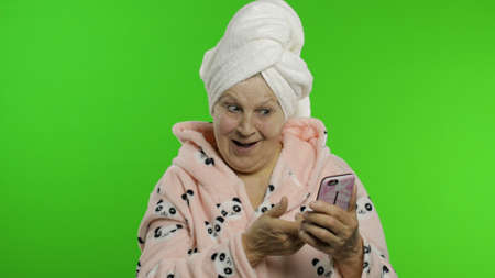 Adult senior caucasian woman grandmother in bathrobe and towel over head using social media app on...