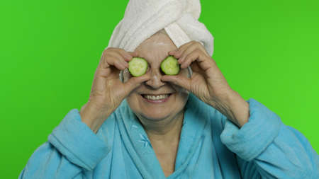 Adult senior caucasian woman grandmother in bathrobe and towel over head with two slices of cucumber applied to eyes isolated on chroma key background. Beauty procedures for mature grandparents peopleの写真素材