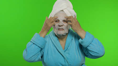 Adult senior caucasian woman grandmother in bathrobe and towel over head applying cosmetic fabric face mask. Chroma key background. Plastic skin care surgery procedures for mature grandparents peopleの写真素材