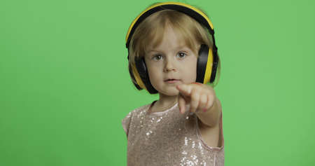 Girl in glossy dress and headphone listening to music and shows a finger forward. Happy four years old girl. Pretty little blonde child. Make faces and smile. Green screen. Chroma Keyの写真素材