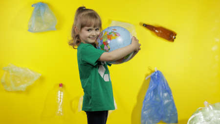 Child girl volunteer in green t-shirt with recycle logo hugs Earth globe. Free from plastic. Yellow background with cellophane bags, bottles. Reduce trash pollution. Save ecology environmentの写真素材