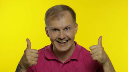 Portrait of young caucasian man 30s posing in pink t-shirt isolated on yellow background in studio. Smiling handsome guy show thumbs up, looking to camera. People emotions, lifestyle conceptの写真素材