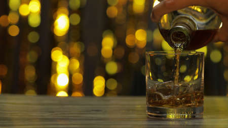 Barman pouring golden whiskey, cognac or brandy from bottle into glass with ice cubes on wooden...
