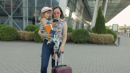 Mother and little daughter stay outdoors near international airport terminal with suitcase luggage on wheels. Woman hold passports and tickets in hand. Child girl and mom tourism, vacation conceptの写真素材