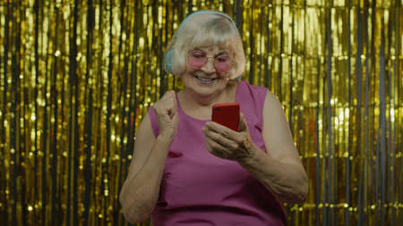 Stylish happy senior old woman in pink blouse holding cell phone, listening to energetic music in...