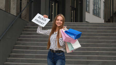 Cheerful trendy girl showing Black Friday inscription text advertisement. Online shopping with low prices. Rejoicing with holiday sale purchase discounts. Shopping mall stairs backgroundの写真素材