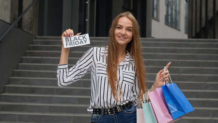Cheerful trendy girl showing Black Friday inscription text advertisement. Online shopping with low prices. Rejoicing with holiday sale purchase discounts. Shopping mall stairs backgroundの写真素材