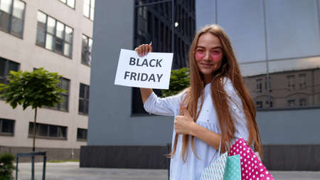 Cheerful trendy girl in short dress showing Black Friday inscription text advertisement. Online shopping with low prices. Rejoicing with holiday sale purchase discounts. Shopping mall backgroundの写真素材