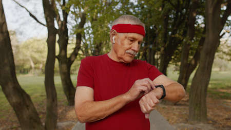 Senior sport runner man using smart watch, tracking distance, checking pulse after fitness workout. Cardio outside in morning park. Active grandparent conceptの写真素材
