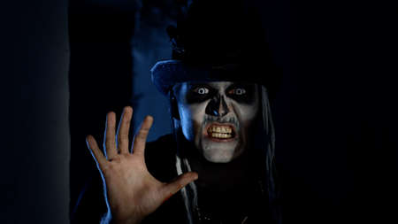 Frightening man with horrible Halloween skeleton makeup in costume with top-hat appears from dark...