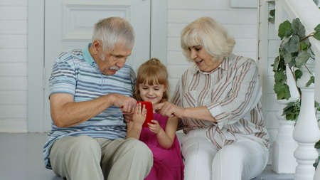 Senior elderly caucasian couple sitting with granddaughter and using mobile phone in porch at home....