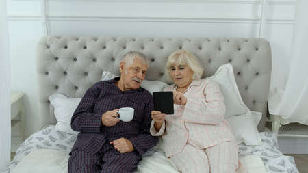 Senior elderly couple wearing pyjamas lying on bed looking on digital tablet making online shopping....