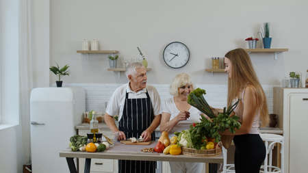 Senior couple in kitchen. Grandparents receiving vegetables from granddaughter. Listening advices of healthy food nutrition. Raw food eating diet. Joyful elderly lifestyle, World vegan day conceptの写真素材