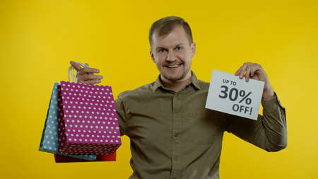 Cheerful man showing Up To 30 percent Off inscription sign and shopping bags, smiling, looking satisfied with low prices, shopping on big sale day. Yellow backgroundの写真素材