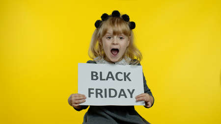 Cheerful stylish blonde child girl in school uniform smiling, holding Black Friday text inscription, rejoicing good discounts, low prices for online shopping sales on holidayの写真素材