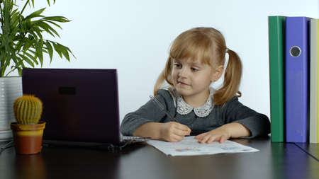 Child schoolgirl learns lessons at home sitting at table using digital laptop computer. Portrait of kid girl distance education. Studying during coronavirus quarantine. White backgroundの写真素材