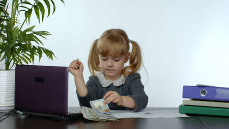 Child girl boss counting dollar cash bills. Baby businesswoman kid with money is sitting at office desk. Finance and business. Lottery online game concept. White backgroundの写真素材