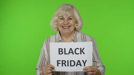 Elderly stylish grandmother smiling and holding Black Friday text inscription banner. Senior woman...