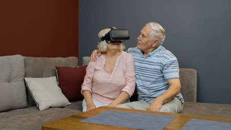 Senior wife playing game in VR headset glasses, husband laughing with her action, elderly couple...