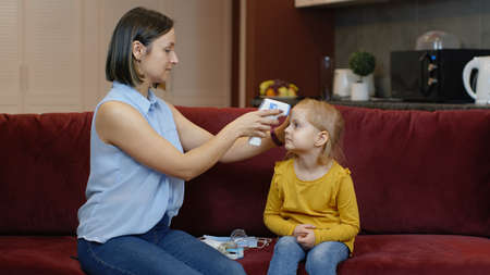 Mother measures temperature with contactless digital thermometer of her little sick daughter child girl at home. Woman with non-contact infrared thermometer. Coronavirus, Covid-19 quarantine lockdownの写真素材