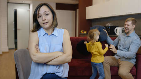 Tired depressed young caucasian mother woman feel very annoyed, suffering migraine of active father and daughter child. Naughty girl jumps, plays noisily with man at home. Hyperractive childrenの写真素材