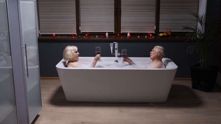 Sexy senior couple grandfather and grandmother is taking foamy bath, drinking red wine in luxury bathroom with candles. Elderly man and woman in love. Spa procedures in hotel. Honeymoonの写真素材