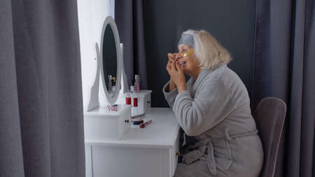 Old senior woman grandmother taking care of skin near eyes and wrinkles, putting makeup on, looking into a mirror. Elderly grandma doing make-up procedures at home. Cosmetics for retired pensionersの写真素材
