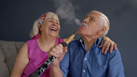 Modern trendy elderly couple smoking hookah at home. Senior grandmother and grandfather having fun relaxing enjoying relationship celebrating birthday. Life of stylish retirees pensionersの写真素材