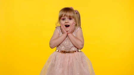 Portrait of happy amazed astonished young kid girl in shiny dress isolated on yellow copy space background. Impressed child. Perfect surprise. Exciting news. Wow reaction.の写真素材