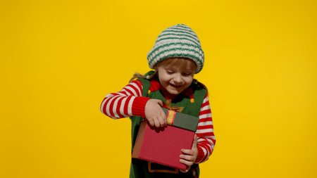 Teen kid girl in Christmas elf Santa helper costume on yellow background. Child getting present gift box, expressing amazement, extreme happiness looking inside box. New Year holidays celebrationの写真素材