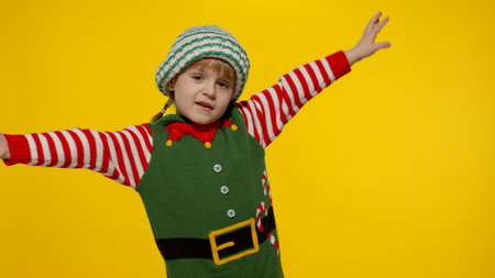 Happy little fun blonde kid teen teenager girl in Christmas elf Santa helper costume dancing, fooling around. Overjoyed child posing isolated on yellow studio background. New Year holidays celebrationの写真素材