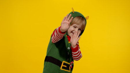 Scared blonde kid teen girl in Christmas elf Santa helper costume looking at camera afraid cover hiding face with hands. Child covering face with fearful expression hides from something terrifyingの写真素材