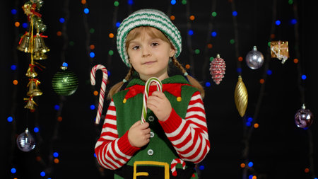 Blonde kid girl in Christmas elf Santa Claus helper costume licking candy cane lollipop isolated on...