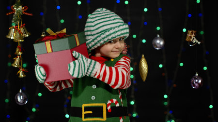 Kid child in Christmas elf Santa Claus helper costume holding present surprise red gift box with ribbon isolated on black background. People New Year holidays celebrationの写真素材