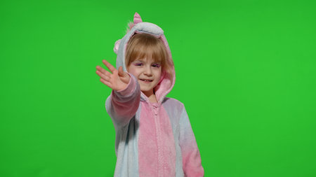 Hello or bye. Little smiling blonde child girl 5-6 years old in unicorn costume posing waving greeting with hand isolated on chroma key green background. Kid animator in unicorn pajamasの写真素材