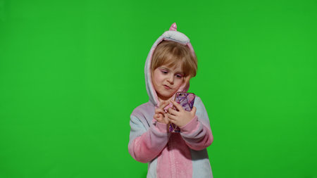 Excited little child girl in unicorn pajamas using, browzing on mobile cell phone, found out great big win news. Kid doing winner gesture isolated on chroma key background. Technology for childrenの写真素材