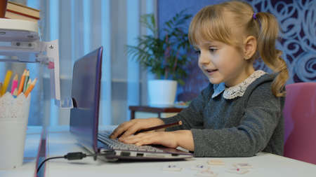 Little child girl doing school homework using laptop at home. Online e-learning, children pupil distance education lesson, technology for kids. Typing on computer and writes in notebook. 6k downscaleの写真素材
