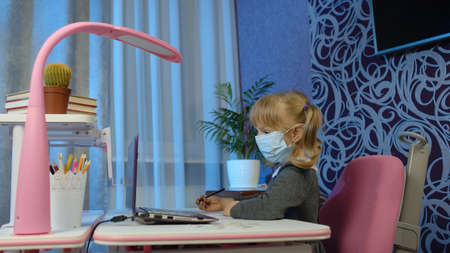 Child girl in mask studying homework with teacher on laptop webcam during online lesson at home....