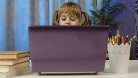 Child schoolgirl learning lessons being at home sitting at table opening laptop digital computer....