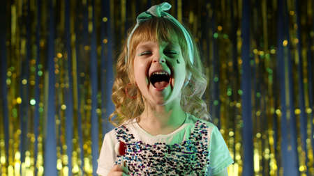 Trendy stylish girl with lollipop candy looking at camera smiling, screaming, shouting. Child kid in neon lights posing at disco club. Futuristic hipster teenager children making faces, fooling aroundの写真素材