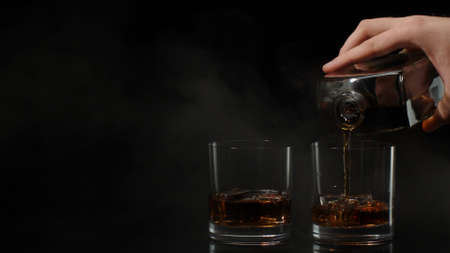Barman pour golden whiskey, cognac or brandy from bottle into glasses with ice cubes on black dark...