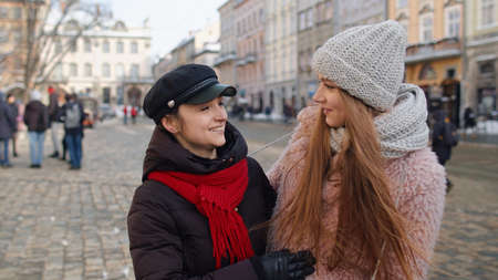 Two smiling women tourists walking together on city street. Sisters couple talking, embracing, having fun, looking at famous sights of old town. Winter holiday traveling. Concept of friends and familyの写真素材