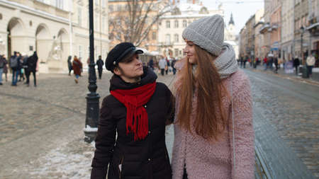 Two smiling women tourists walking together on city street. Family couple talking, embracing, having...