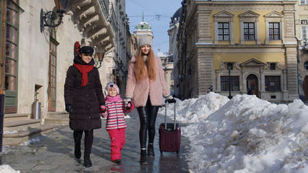 Two young smiling women travelers with adoption child girl walking with suitcase, looking at famous sights of old city streets. Lesbian couple talking, smiling outdoor. Winter holidays vacation tripの写真素材